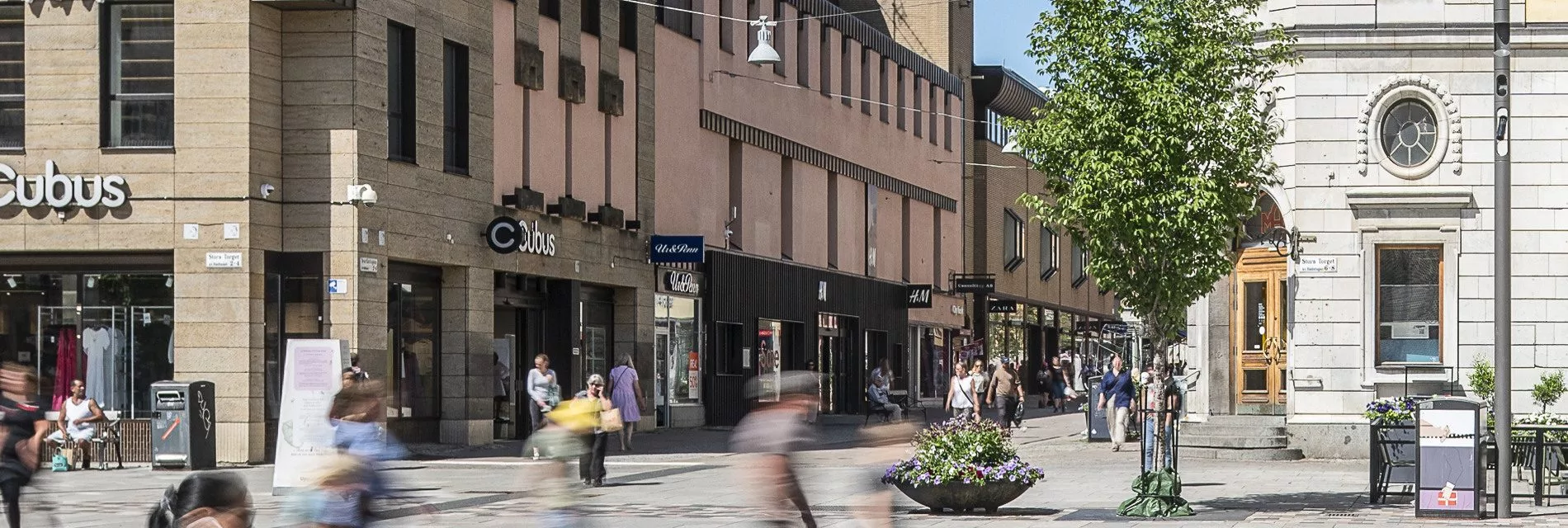 En stadsgata med människor som går förbi butiker, där fasaderna har en rosa färg och blomdekorationer syns.