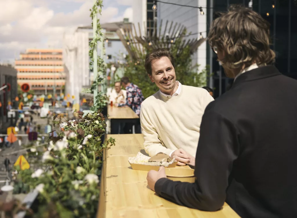 Två personer pratar vid ett bord utomhus på en solig dag, med blommor och byggnader i bakgrunden.