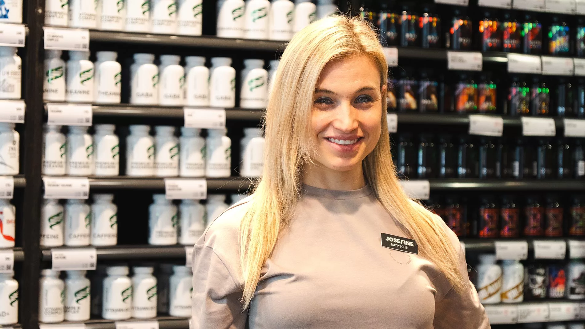 En kvinna med långt blont hår står framför hyllor fyllda med burkar och produkter.
