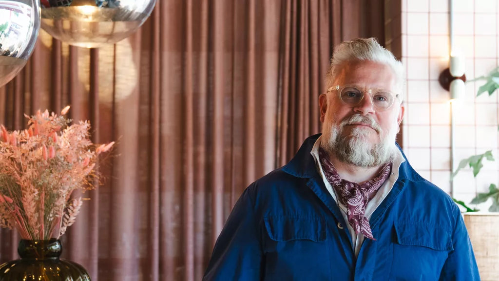 En man med grått skägg och glasögon, iklädd en blå jacka och en halsduk, står framför bruna gardiner.