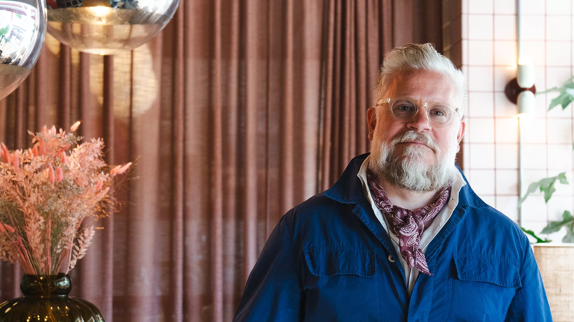 En man med grått skägg och glasögon, iklädd en blå jacka och en halsduk, står framför bruna gardiner.