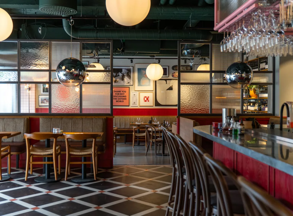 En restauranginteriör med svartvitt golv, stora hängande lampor, barstolar vid en röd bardisk, och bord vid fönstersidor.