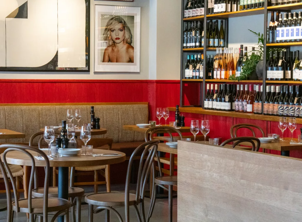 En restaurang med röda väggar, träbord och stolar, samt hyllor fyllda med vinflaskor och en tavla på väggen.