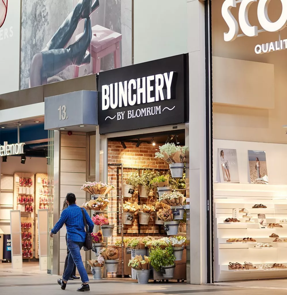 En butik med namnet "Bunchery" har blomsterarrangemang vid entrén, och en person går förbi på trottoaren.