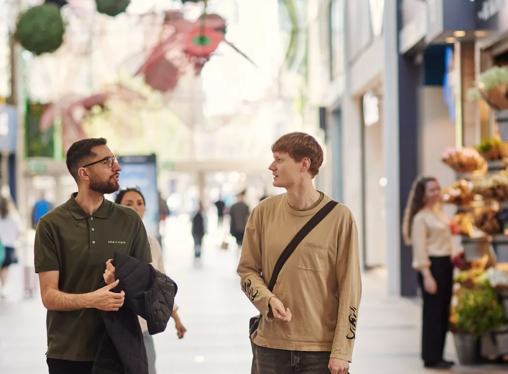 Två män samtalar och går i en galleria med dekorationer och andra besökare i bakgrunden.
