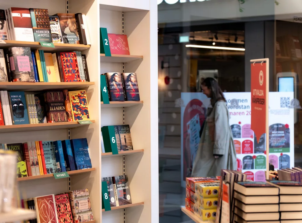 Bokhyllor fyllda med böcker står nära ett butiksfönster, där en person kan ses gå förbi utanför.