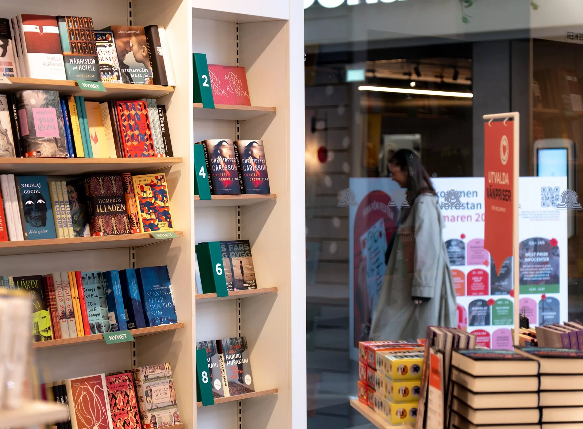 Bokhyllor fyllda med böcker står nära ett butiksfönster, där en person kan ses gå förbi utanför.