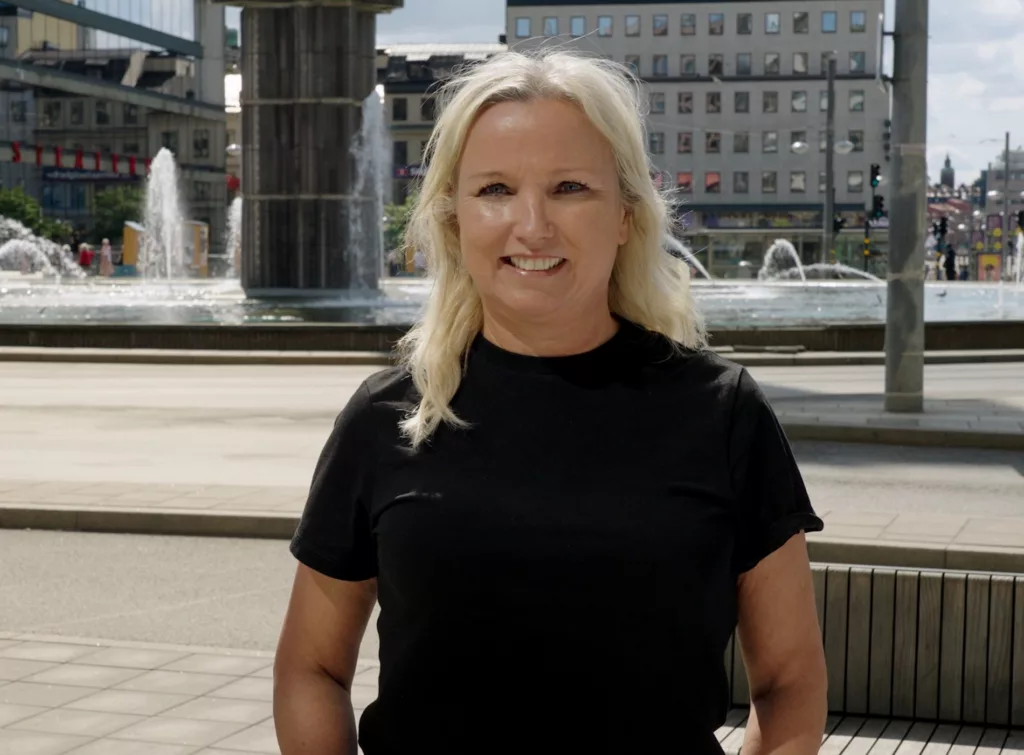 En kvinna med blont hår står framför en fontän och byggnader i en stadsmiljö.