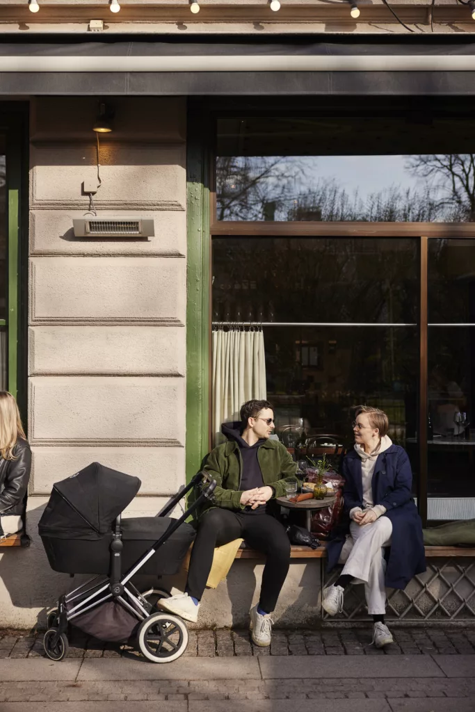 Två personer sitter vid ett cafébord utomhus med en barnvagn bredvid dem, omgivna av grönska och en byggnad.