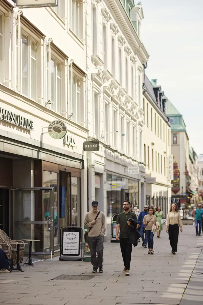 Flera människor promenerar på en shoppinggata med vita byggnader och skyltar längs sidorna.