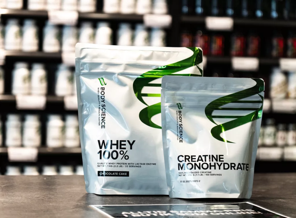 Två förpackningar med texten "Whey 100%" och "Creatine Monohydrate" står på ett bord, omgivna av en butikshylla med produkter i bakgrunden.