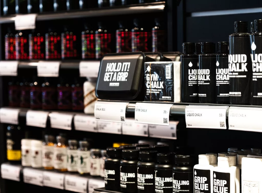 Flera hyllor fyllda med burkar och flaskor med kosttillskott, inklusive en svart förpackning med texten "Hold It Retail" och "Liquid Chalk".