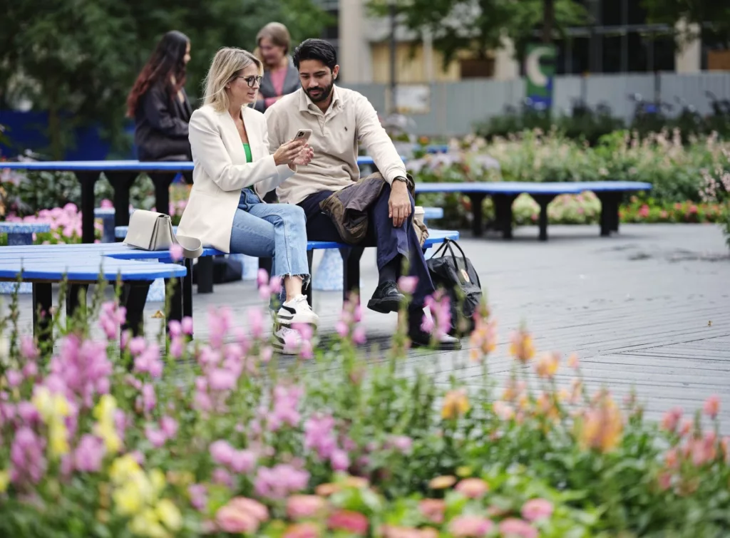 Två personer sitter på en parkbänk och tittar på en mobiltelefon, med blommor och grönska i förgrunden.