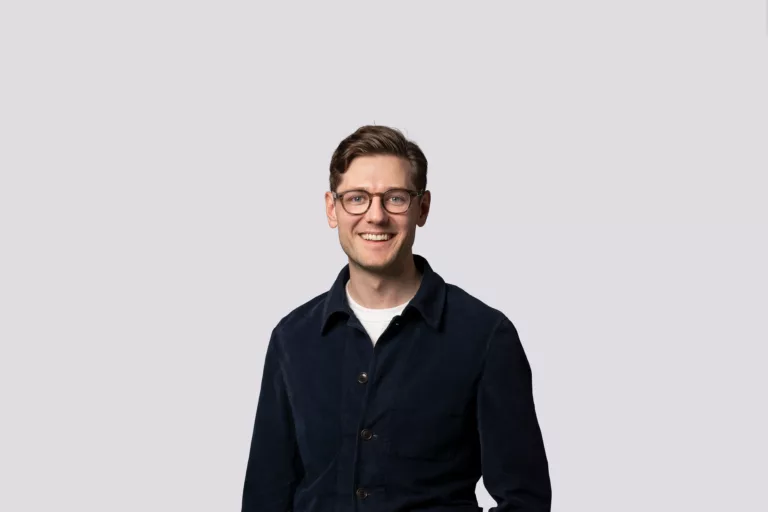 En man med glasögon och kort brunt hår ler, iklädd en mörkblå skjorta och vit t-shirt, mot en ljusgrå bakgrund.
