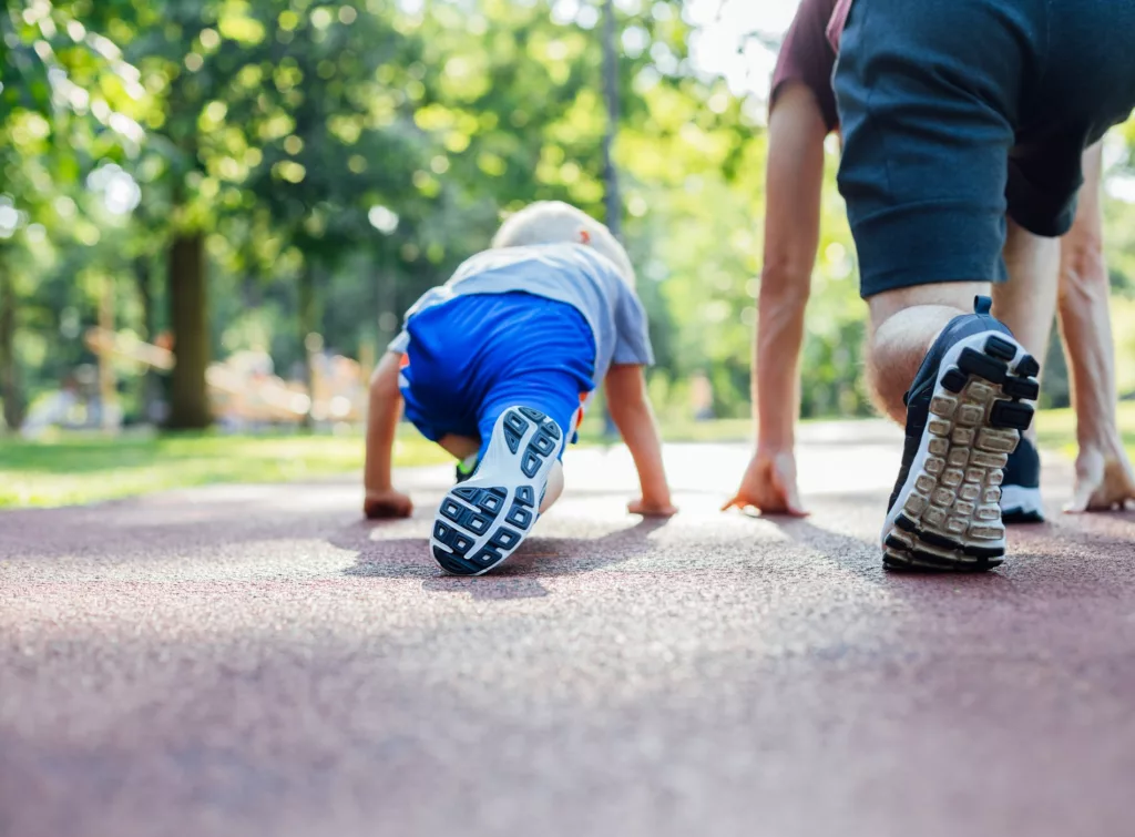 5. Ett barn kryper på en löparbana bredvid en vuxen person, med grönska i bakgrunden.