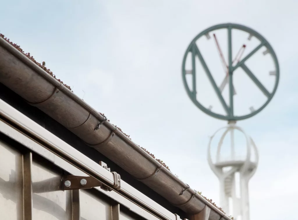 En närbild av ett tak med en ränna i förgrunden och en cirkulär skylt med bokstäverna "NK" på en mast i bakgrunden.