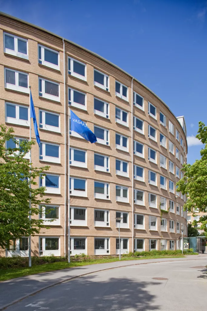 1. En rundad flerbostadsbyggnad i tegel med flera våningar och blå flaggor framför, omgiven av träd under en klar blå himmel.