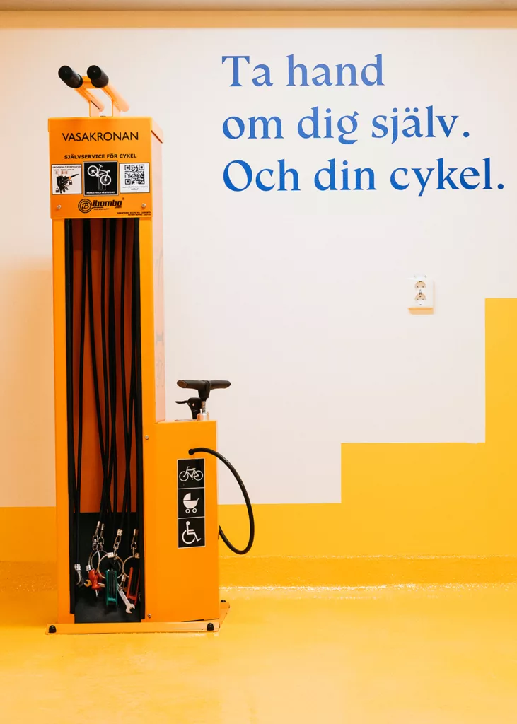 En orange cykelstation med pumpar och verktyg, med texten "Ta hand om dig själv. Och din cykel." på väggen.