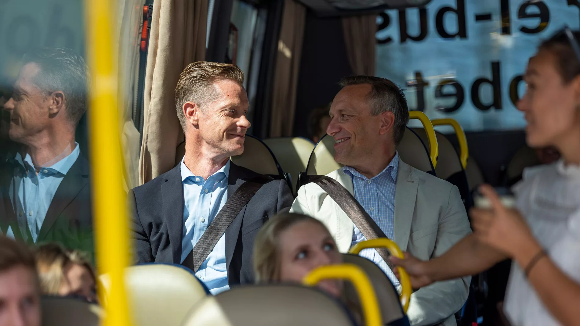 3. Två personer i kostym skrattar och samtalar medan de sitter på en buss. En kvinna syns i förgrunden.