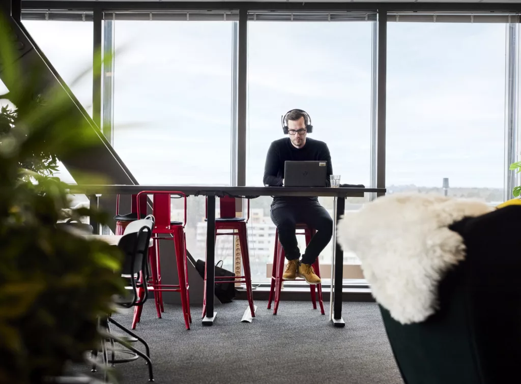 En person med hörlurar sitter vid ett högt bord med röd barstol och arbetar på en laptop nära ett stort fönster.