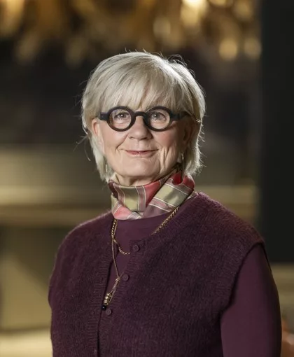 En äldre kvinna med glasögon, klädd i en mörklila tröja och en mönstrad halsduk, står framför en suddig bakgrund.