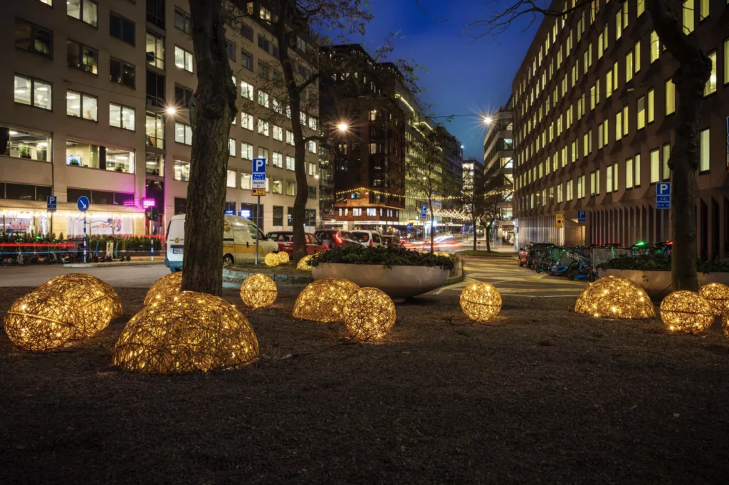 Belysta guldfärgade dekorationer i form av klot placerade i en park, omgivna av höga byggnader och en kvällsupplyst stad.