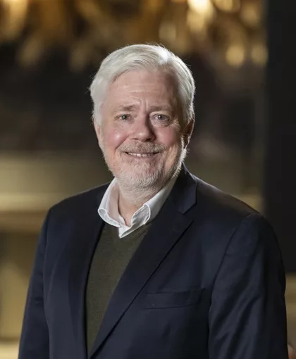 En äldre man med vitt hår och skägg ler, klädd i en mörk kavaj och grön tröja, med en suddig bakgrund.