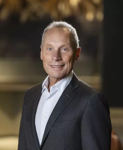 En man med kort grått hår och mörk kostym ler mot kameran. Bakgrunden är suddig och har mörka och varma färger.