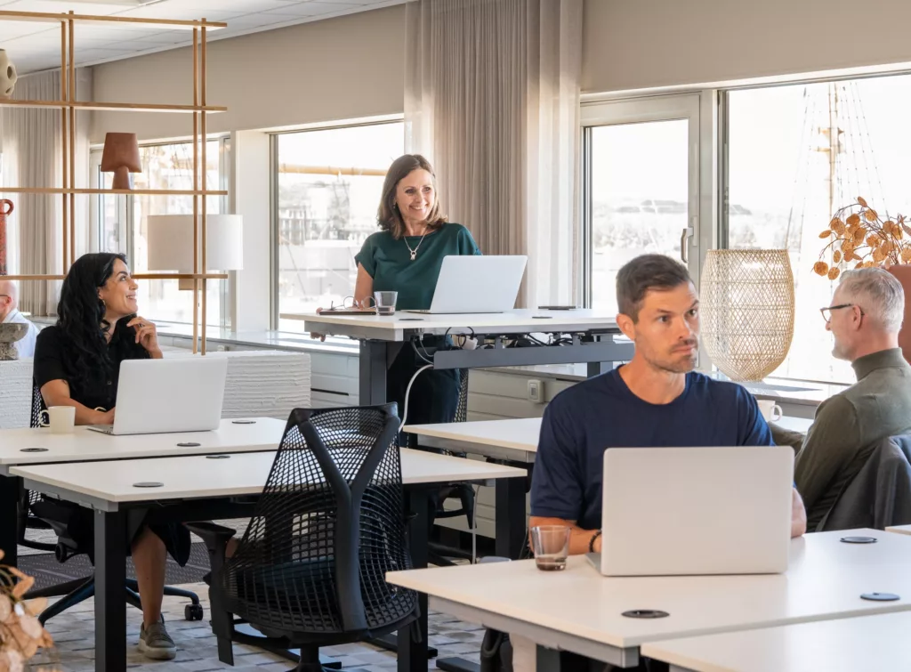 Flera personer arbetar vid skrivbord med laptops i ett ljust och modernt kontorslandskap; en person står vid ett höj- och sänkbart bord.