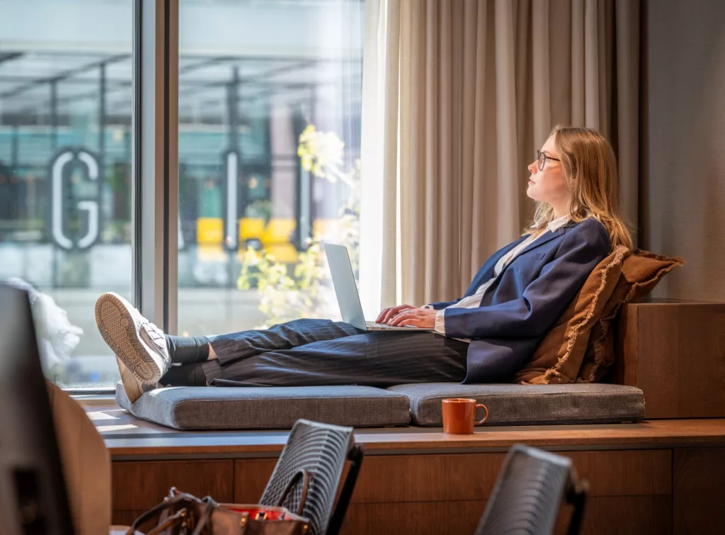 En person sitter avslappnat med benen uppe på en sittmöbel, tittar ut genom ett stort fönster och använder en laptop.