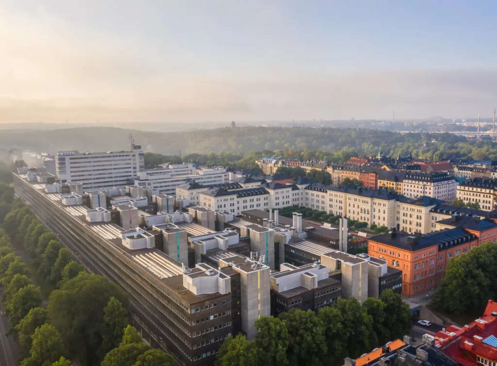 Flygvy över en stadsdel med flera moderna byggnader, träd och en dimmig himmel i bakgrunden.