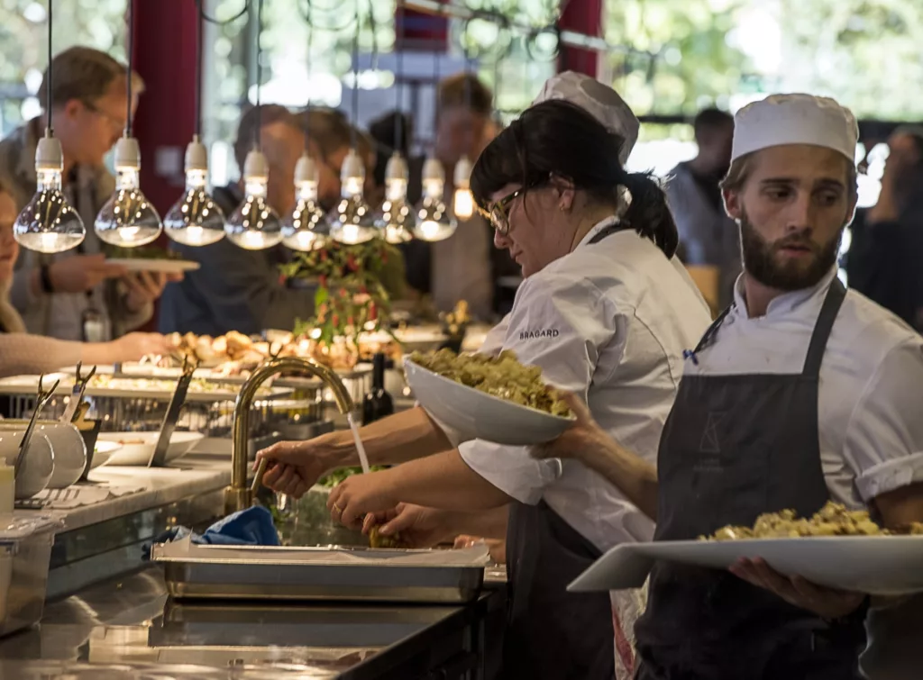 Kockar i arbetskläder förbereder mat och serverar tallrikar i ett restaurangkök med belysning ovanför disken.