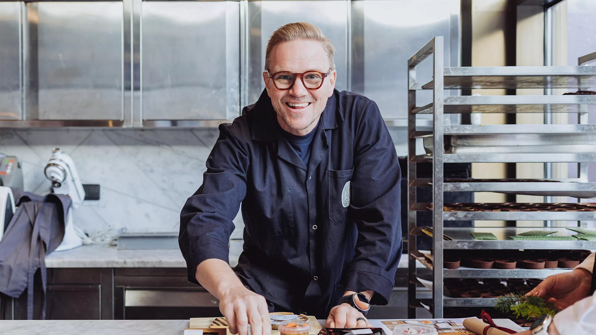 En man med glasögon och mörk arbetsrock ler och arbetar vid ett bord i ett professionellt kök.