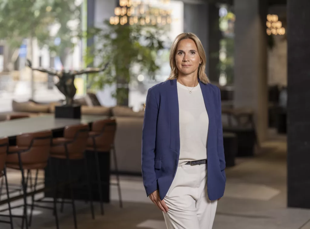 En kvinna klädd i en blå kavaj och vit topp står i en ljus och modern inomhusmiljö med gröna växter och möbler i bakgrunden.