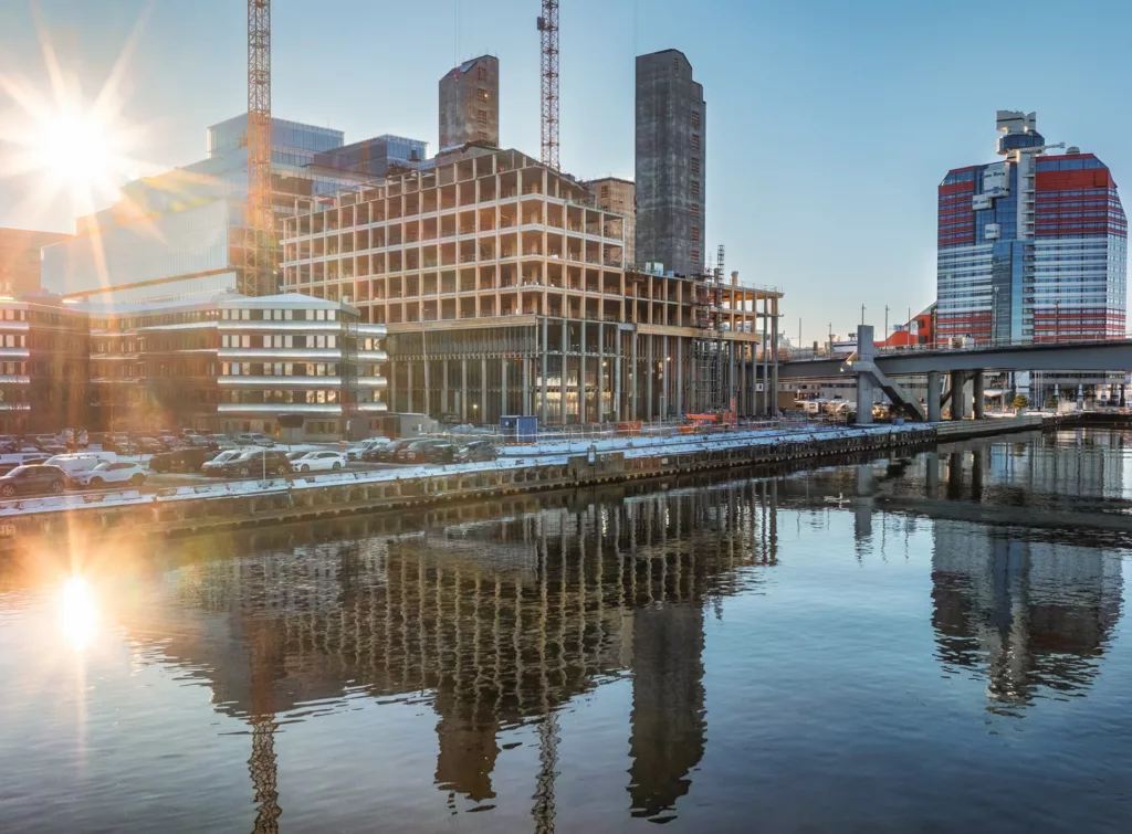 2. En byggarbetsplats vid vattnet med höga byggnader, kranar och reflektioner i vattnet under en solig himmel.