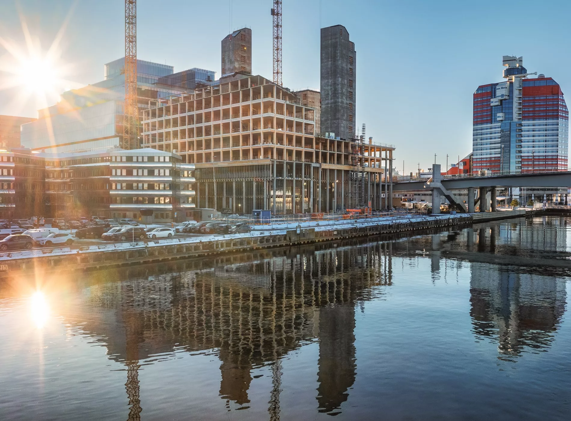 2. En byggarbetsplats vid vattnet med höga byggnader, kranar och reflektioner i vattnet under en solig himmel.