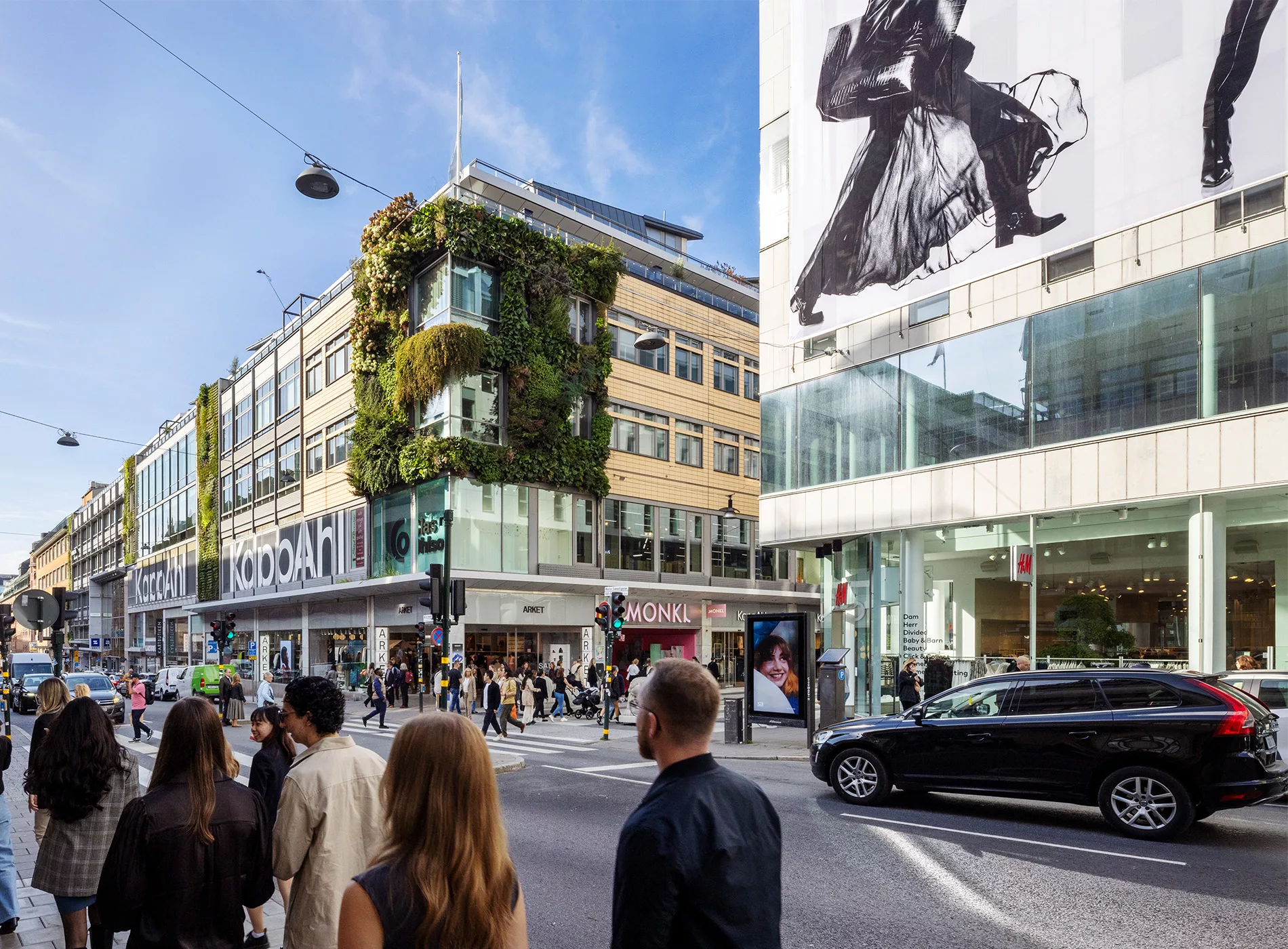 En stadskorsning med människor som går, en byggnad täckt av grönska på ena hörnet, och en reklam på en fasad med en modell i svart klädsel.
