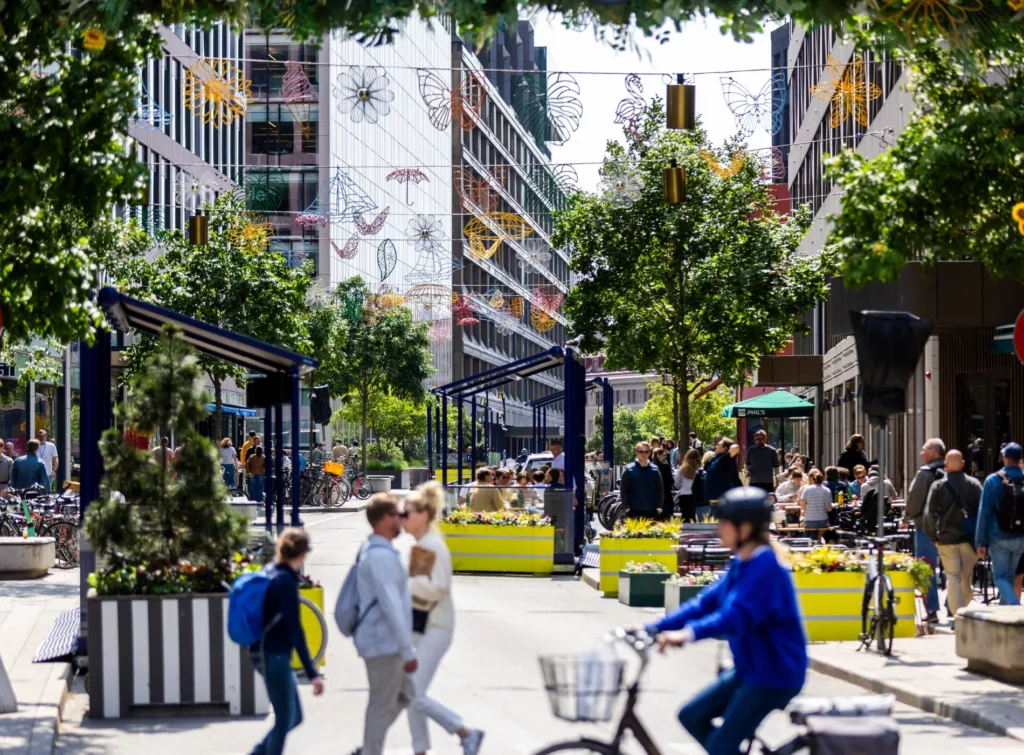 4. En stadsmiljö med människor som promenerar och cyklar bland moderna byggnader, träd och dekorationer.