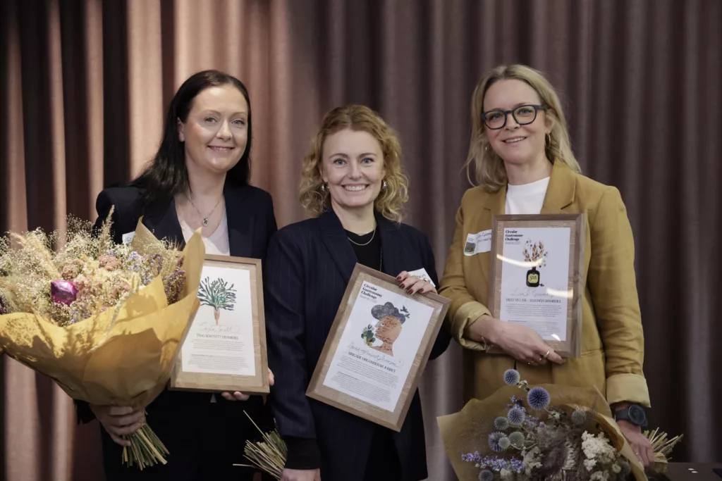 Tre kvinnor poserar med blommor och inramade diplom framför ett draperi.