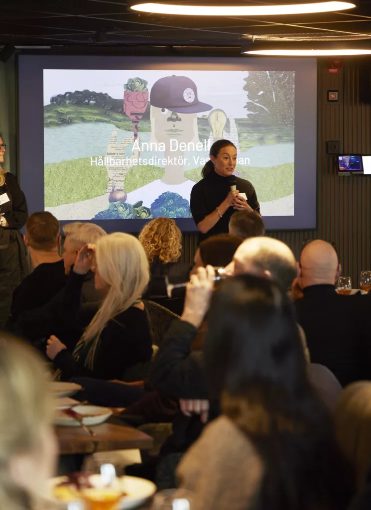 En kvinna står och talar framför en publik, med en presentation projicerad på en skärm i bakgrunden.