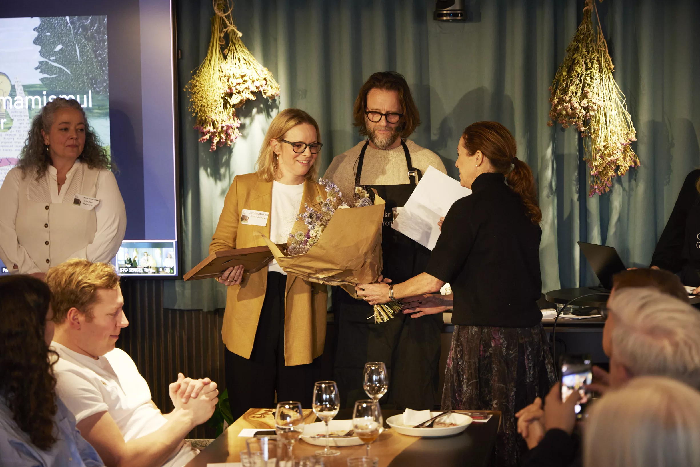 En kvinna överlämnar en bukett blommor och dokument till två personer på en scen i en restaurangmiljö.