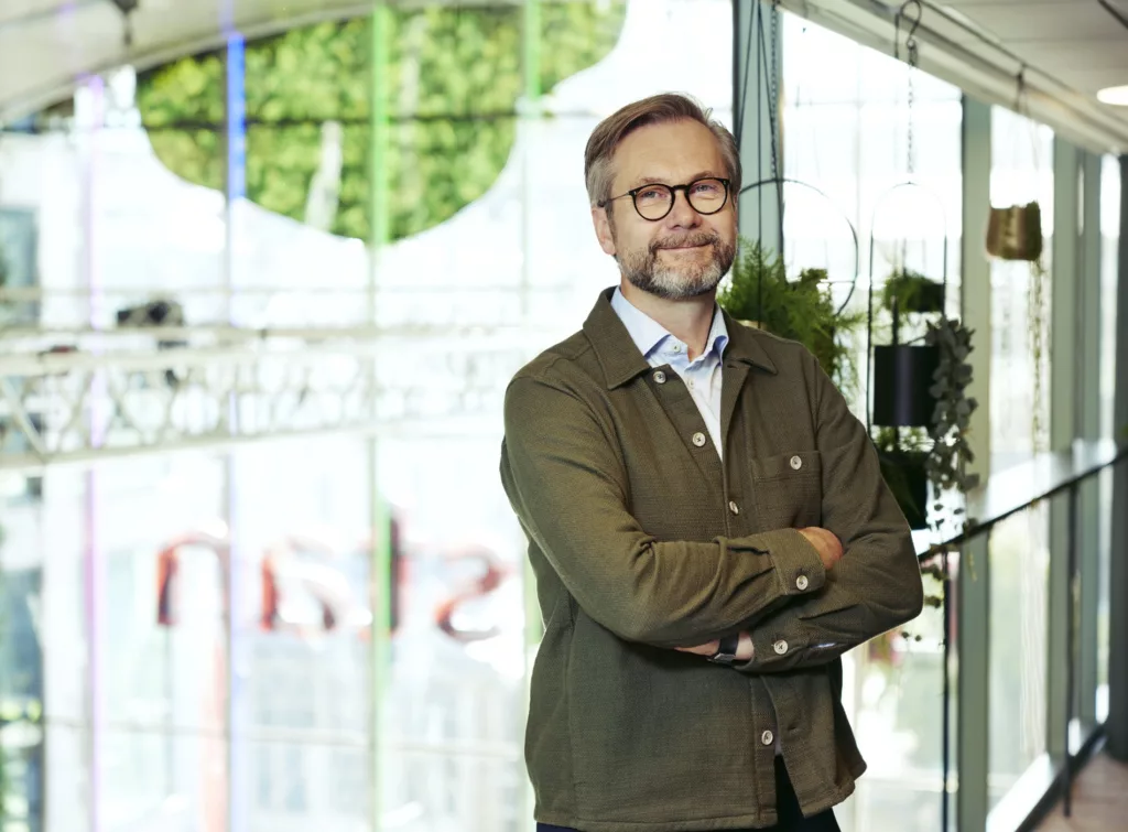 En man med glasögon och skägg står leende med armarna i kors framför en ljus och grönskande bakgrund.