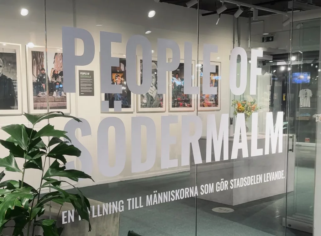 Glasvägg med texten "PEOPLE OF SÖDERMALM" och en växt i förgrunden.