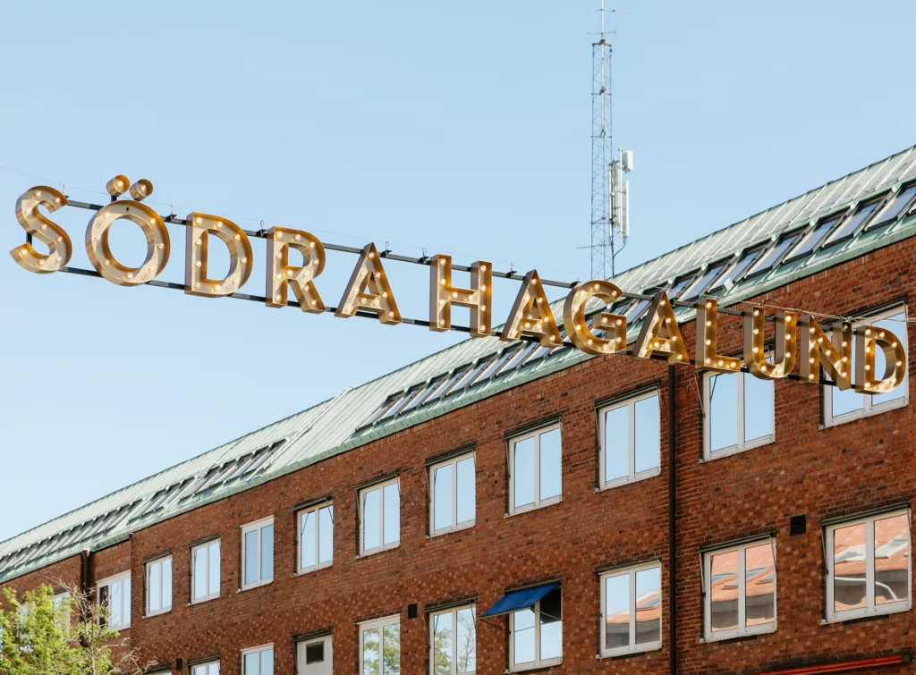 1. Texten "Södra Hagalund" i guldbokstäver hänger ovanför en röd tegelbyggnad under en klarblå himmel.