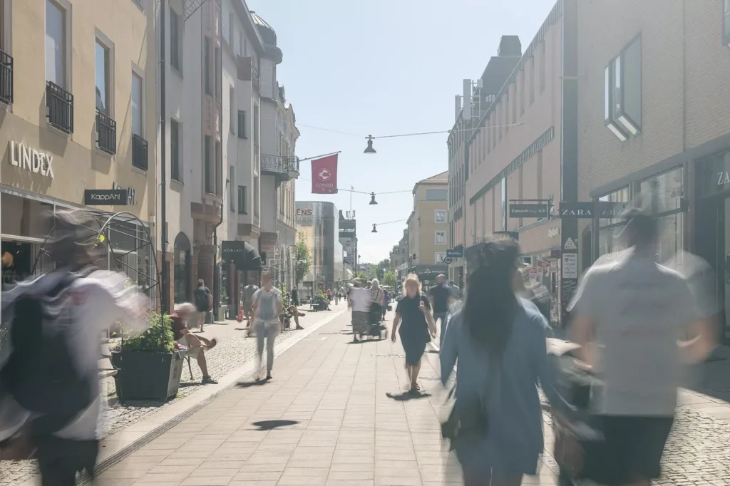 En solig stadsgata med människor som promenerar och flaggor hänger ovanför.