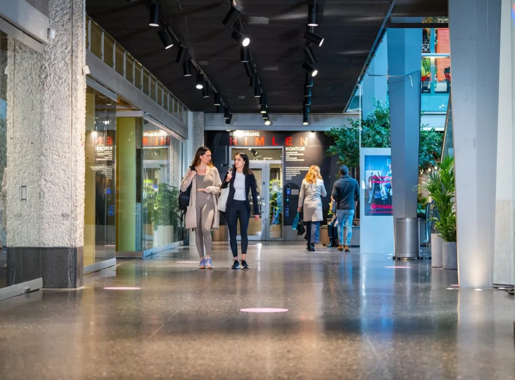 Två kvinnor går genom en modern shoppinggalleria med glasfasader och dekorativa växter.