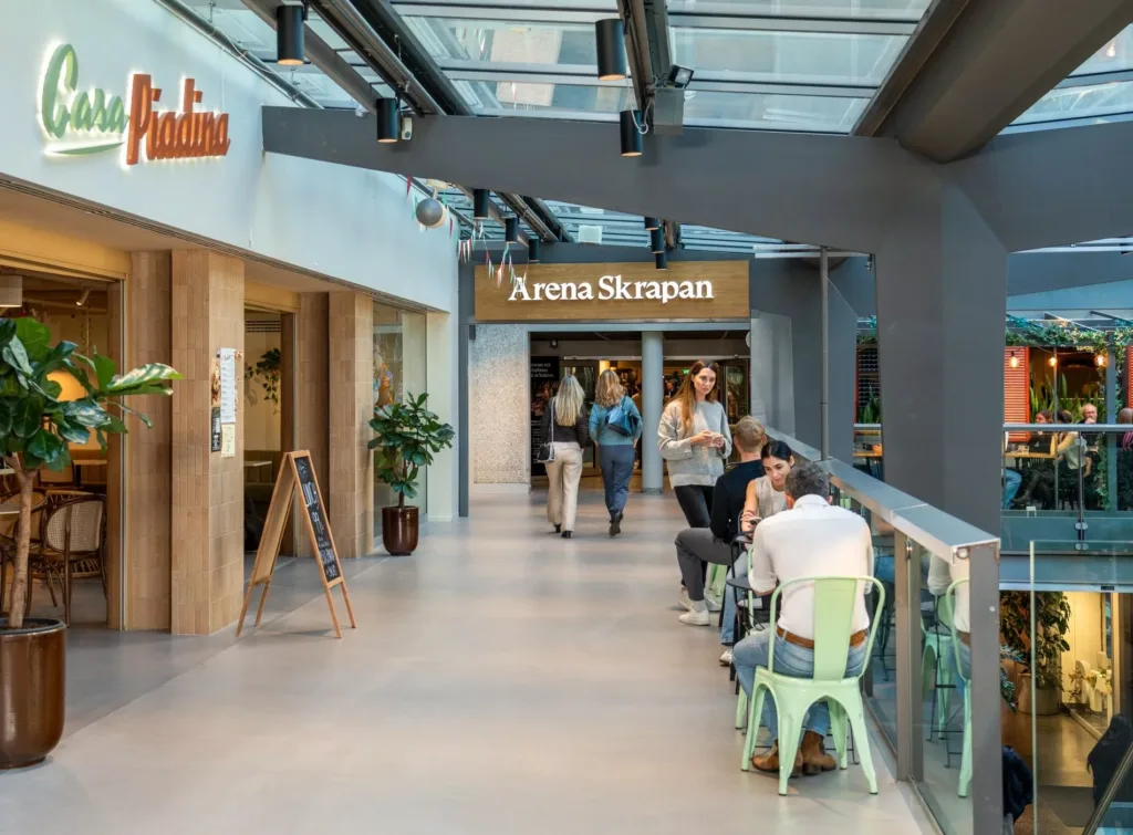 En korridor med glasväggar, människor som promenerar och skyltar för restauranger, inklusive "Arena Skövde".