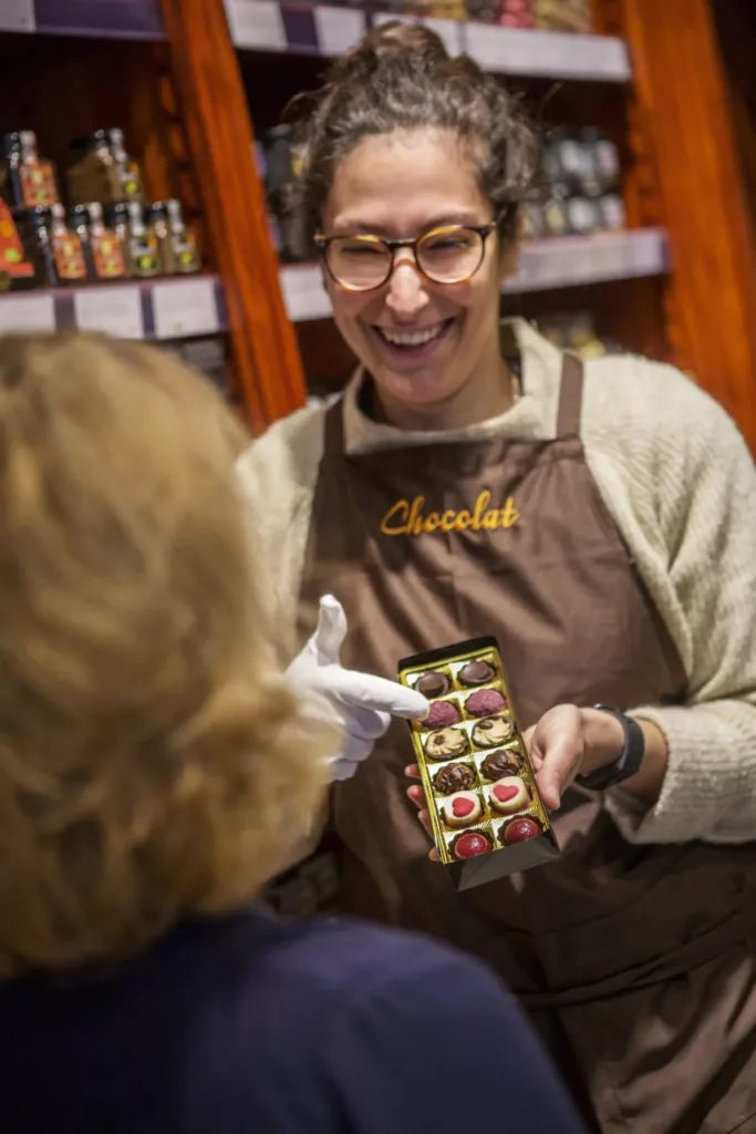 En leende person i ett förkläde med texten "chocolat" visar en ask med färgglada praliner för en kund.