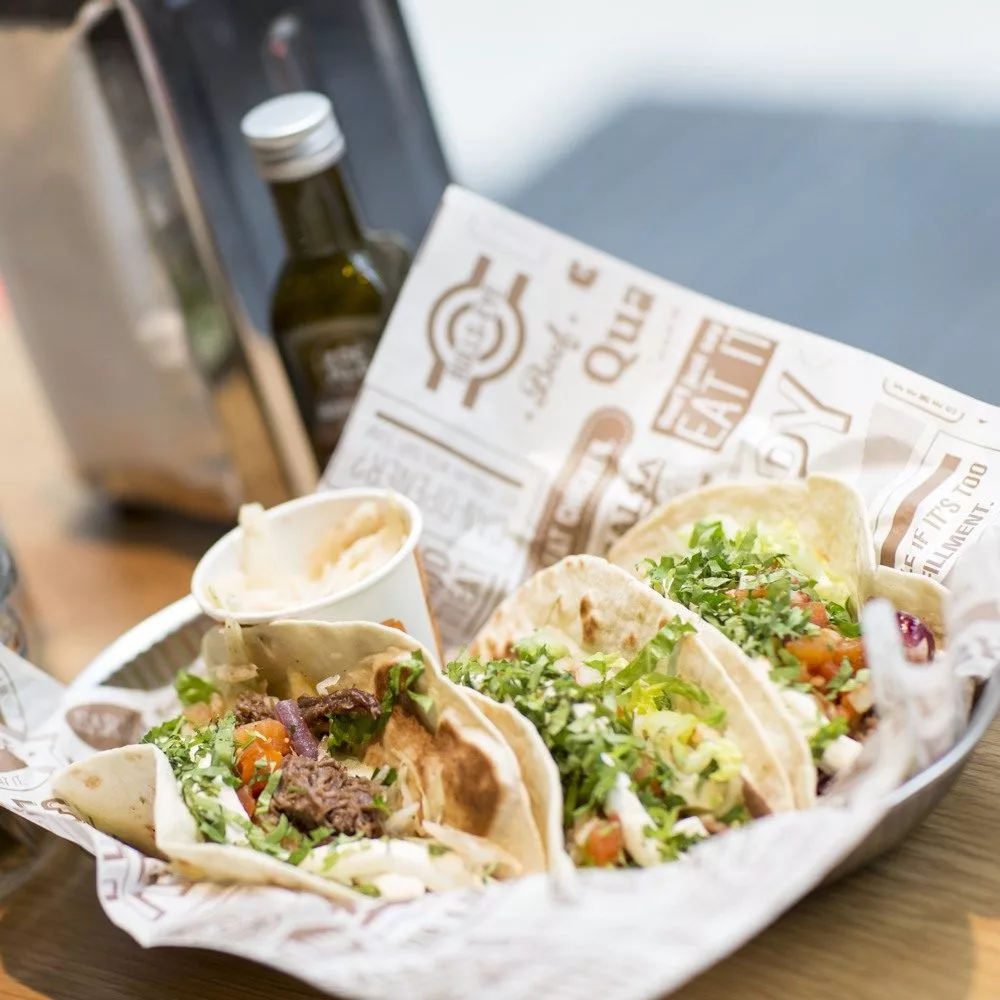 Tre tacos fyllda med kött, grönsaker och sås serverade i en korg med papper, en liten såskopp och en flaska olivolja bredvid.