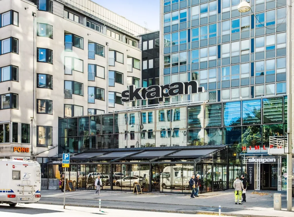 En byggnad med glasfasad och texten "Skrapan" på toppen, omgiven av stadsmiljö.
